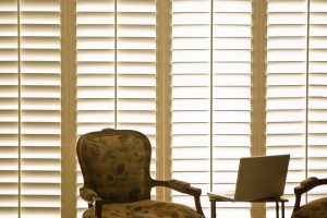 Armchair sitting in front of a laptop with shutters behind | Featured image for Pros and Cons of PVC Shutters Blog from U Blinds Australia.