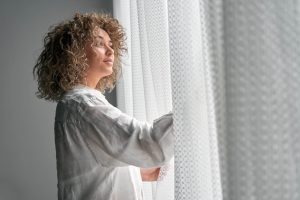 Woman in a white t-shirt opening white curtains | Featured image for How Energy Efficient Window Coverings Can Save You Money Blog by U Blinds Australia