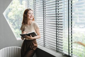 Woman sitting on a window sill and smiling while looking outside | Featured image the Inside Mount vs Outside Mount – Knowing Which to Choose Blog by U Blinds Australia.