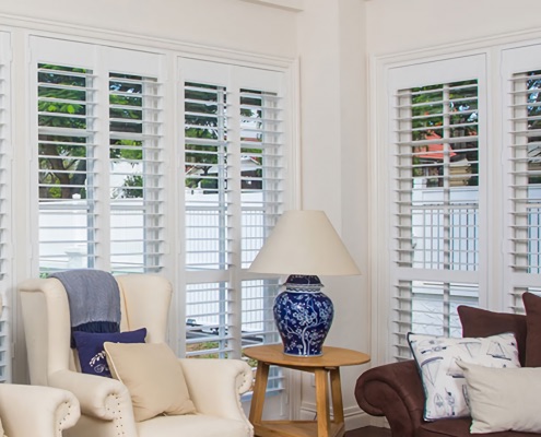 Modern living room with white walls and white PVC plantation shutters and a blue lamp on a small wood table | Featured Image for the PVC Plantation Shutters Page of U Blinds Australia.