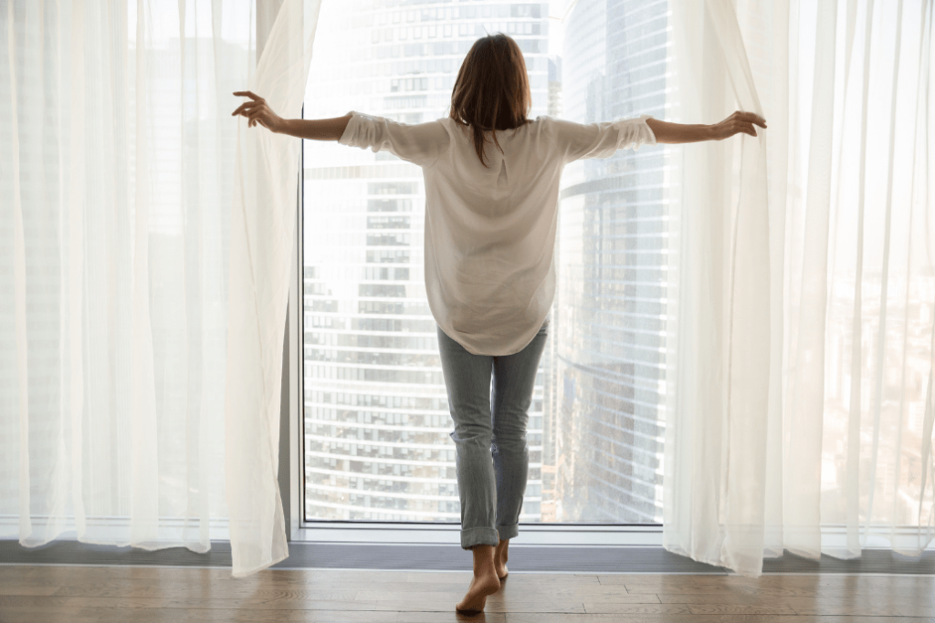 Woman opening curtains Hobart adores and looking out over a city | Featured image for the Blinds Hobart location page by U Blinds Australia.